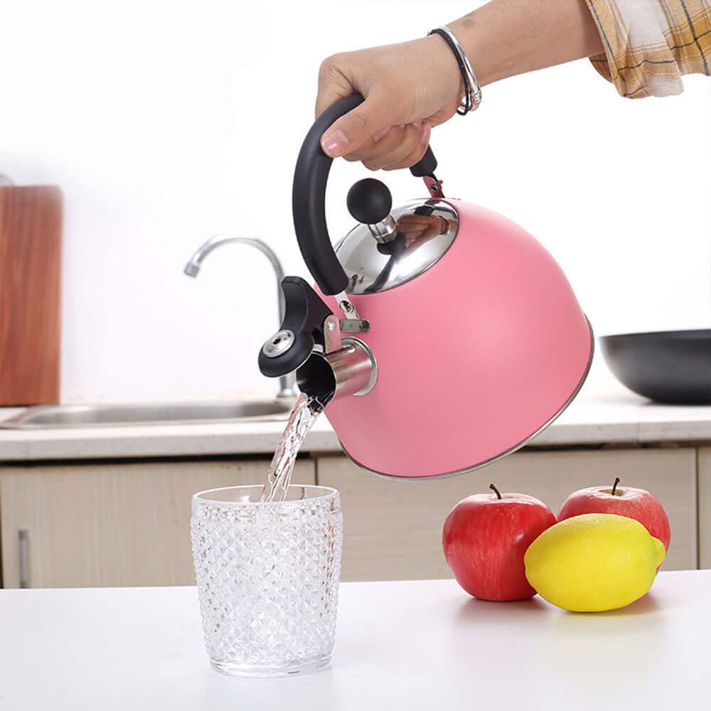 Pink stainless steel whistling kettle pouring water into a glass with apples and lemon on the kitchen counter.