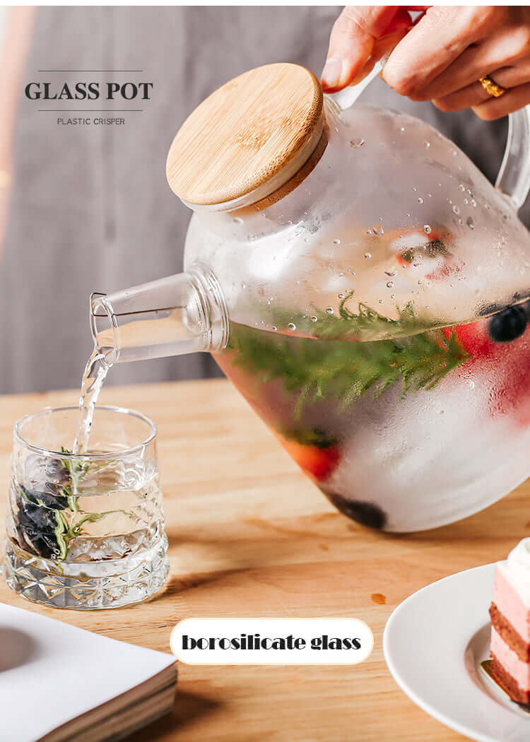 Nordic glass teapot with bamboo lid pouring infused drink into a glass, showcasing fresh fruits and herbs.