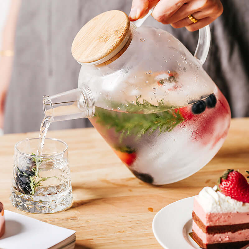 Nordic style transparent kettle with bamboo lid pouring flavored water into a glass, showcasing fresh ingredients.