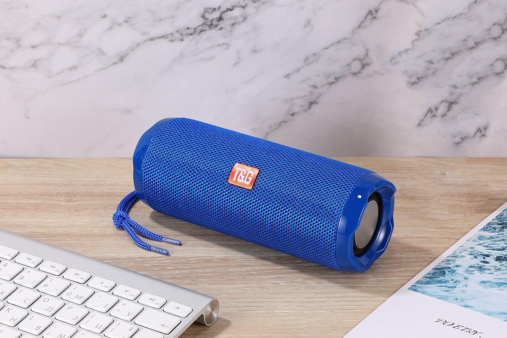 Blue portable Bluetooth speaker with card holder on a wooden desk next to a keyboard and a photo.