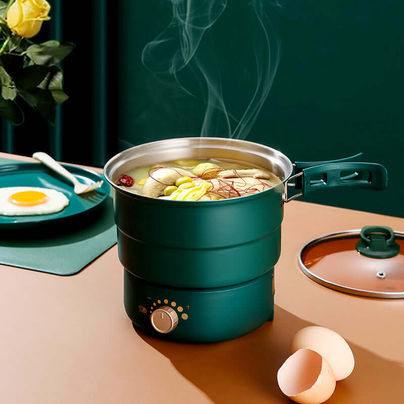 Electric cooker with steaming vegetables, eggs, and a glass cover on a table with plates and a green background.