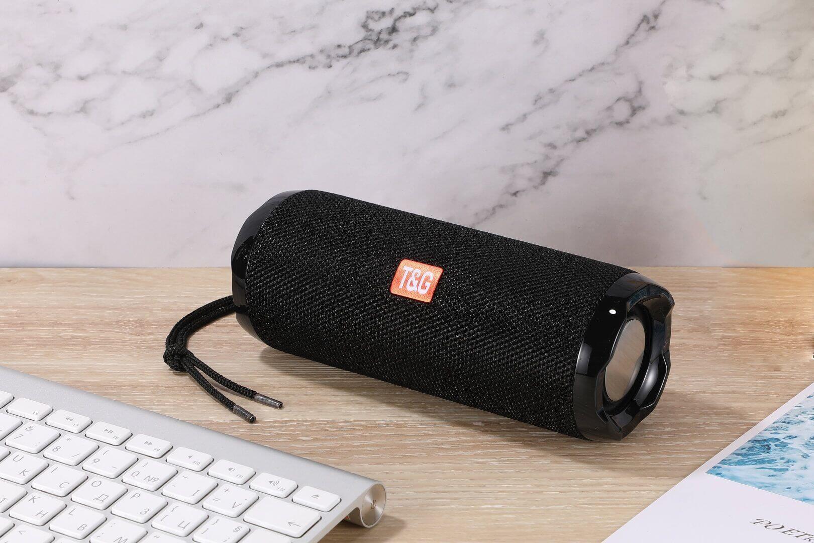 Black fabric portable Bluetooth speaker with card holder next to a keyboard on a wooden table.