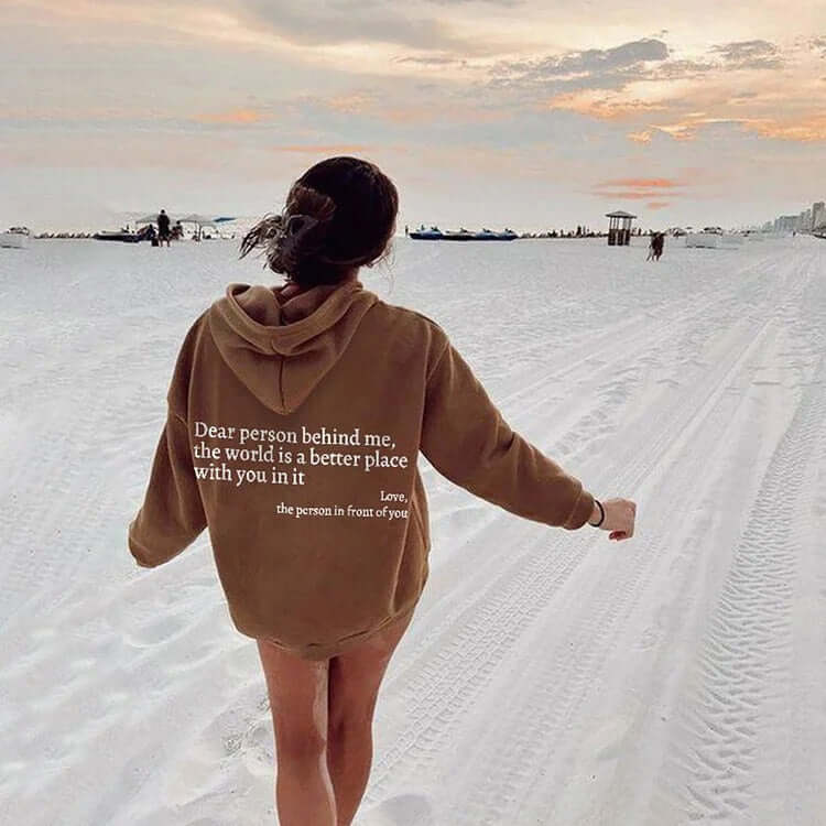Woman walking on the beach wearing a brown hoodie with the quote "Dear person behind me, the world is a better place with you in it"