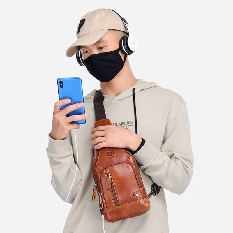 Casual style individual wearing a brown leather messenger bag while using a smartphone and wearing headphones.