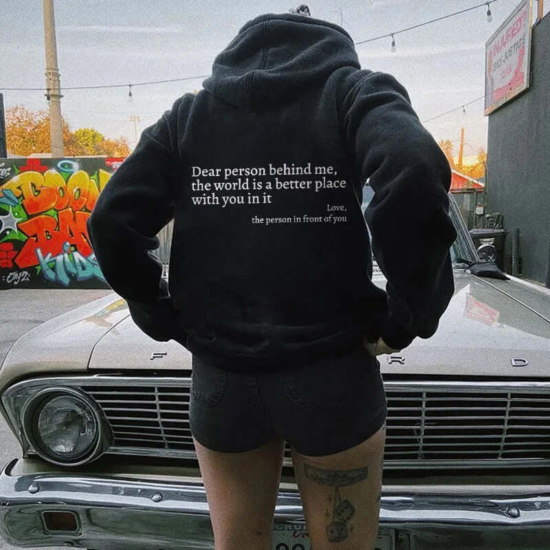 Person wearing black hoodie with motivational text on back, styled with denim shorts and urban backdrop.