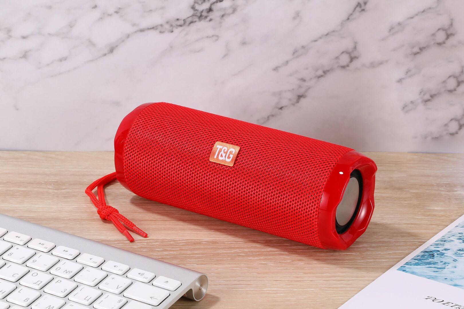 Red portable Bluetooth speaker with card holder on wooden desk beside keyboard and notebook.